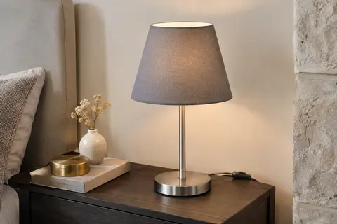 Lampe de chevet à pied en métal nickelé mat et abat-jour en tissu gris, posée sur une table en bois.