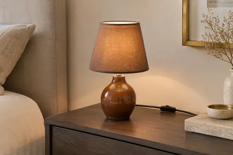 Lampe de chevet à pied en céramique brune et abat-jour en tissu, posée sur une table en bois.