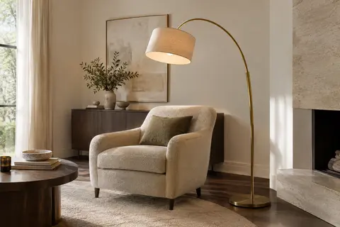 Lampadaire arc doré avec abat-jour en lin beige orienté près d’un fauteuil de salon