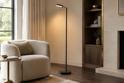 Lampadaire LED noir à structure élancée, installé près d’un fauteuil dans un salon à la lumière chaude.
