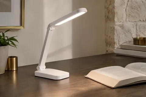 Lampe de bureau blanche pliable et orientable, allumée sur un bureau en bois près d’un livre ouvert